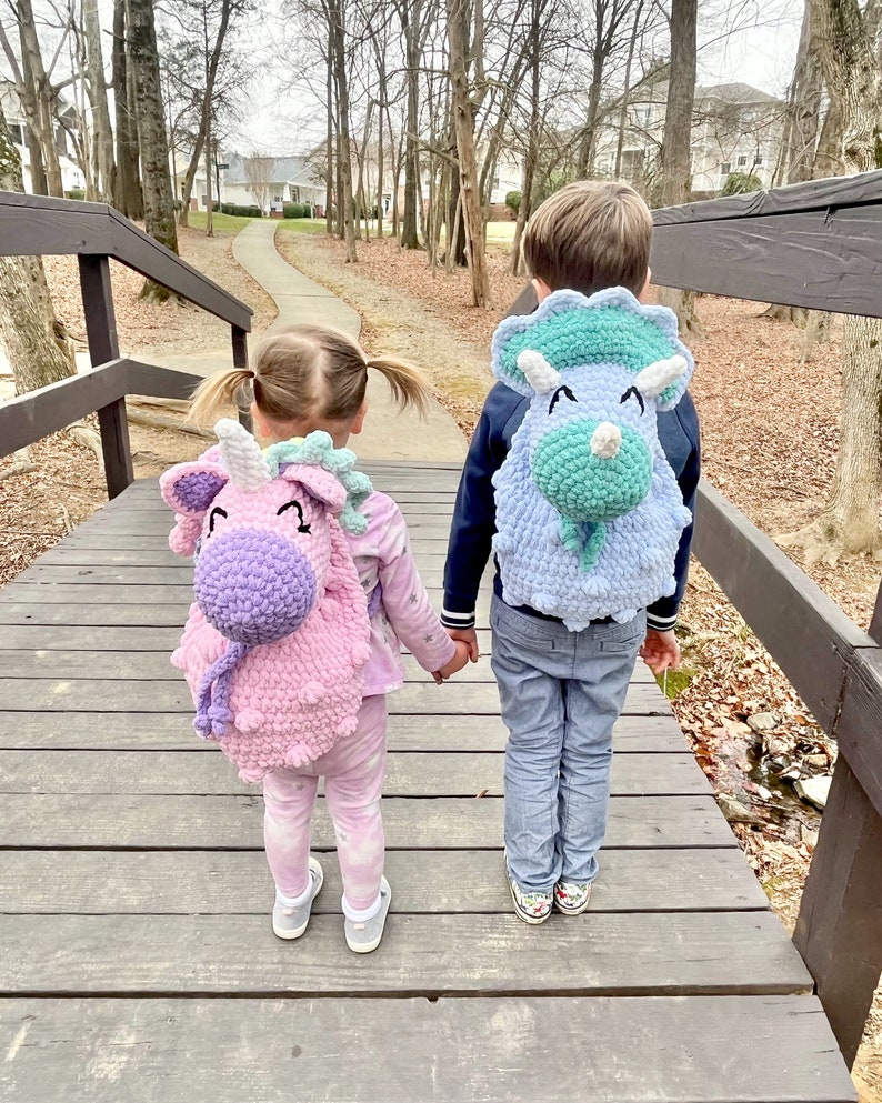 May include: Two children, one wearing a pink unicorn backpack and the other wearing a blue triceratops backpack, walk hand-in-hand on a wooden bridge.