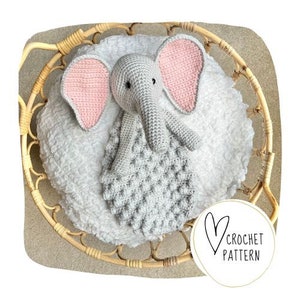 May include: A crocheted elephant baby blanket with pink ears and a gray body. The blanket is sitting in a wicker basket.