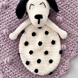 May include: A white crocheted dog with black spots and black ears, lying on a purple crocheted blanket with a textured surface.