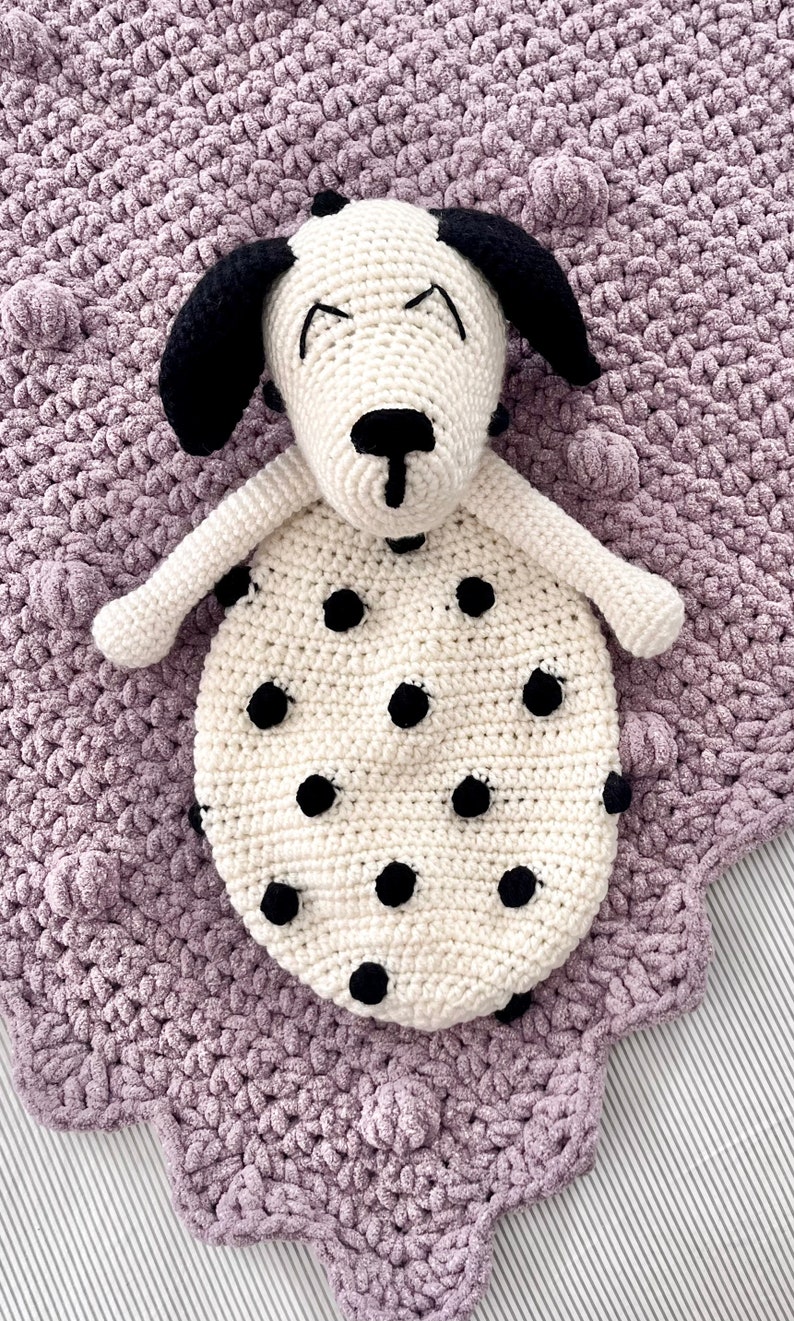 May include: A white crocheted dog with black spots and black ears, lying on a purple crocheted blanket with a textured surface.