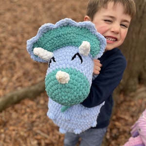 May include: A young child holds a crocheted blue and green dinosaur toy. The dinosaur has white horns and eyes. The child is smiling and looking at the camera.