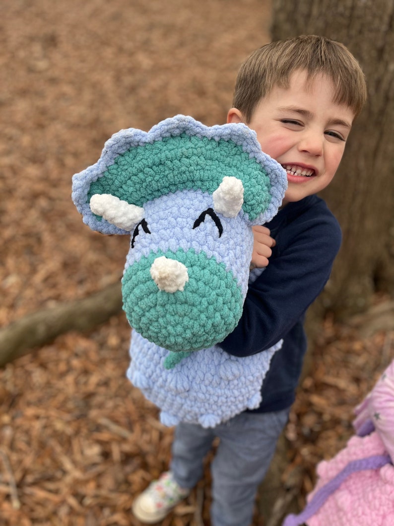 May include: A young child holds a crocheted blue and green dinosaur toy. The dinosaur has white horns and eyes. The child is smiling and looking at the camera.