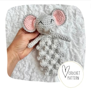 May include: A gray crocheted elephant amigurumi toy with pink ears and a black embroidered face. The toy is lying on a white fluffy surface.