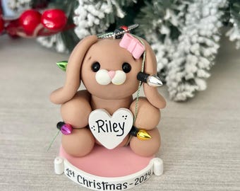 May include: A brown bunny ornament with a pink bow and a heart that says "Riley" on it. The ornament is decorated with Christmas lights and says "1st Christmas - 2024" on the base.