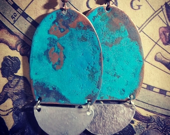 May include: A pair of oval-shaped earrings with a turquoise blue and copper patina finish. The earrings have a hammered silver half-moon charm hanging from the bottom.
