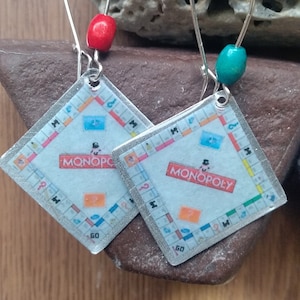 May include: A pair of silver earrings with a Monopoly board game design. The earrings have a silver hook and a red and turquoise bead at the top.