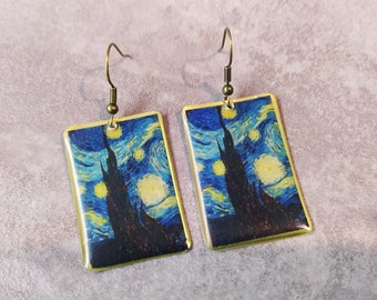 May include: A pair of rectangular earrings featuring a print of Van Gogh's "Starry Night" painting. The earrings have a gold-colored border and a bronze-colored hook. The artwork depicts a dark, tall cypress tree under a swirling blue sky filled with stars.