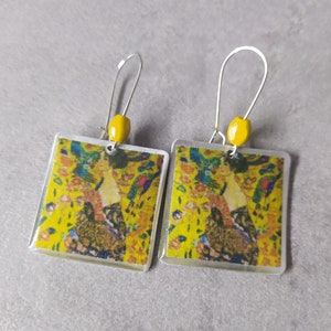 May include: A pair of silver earrings with yellow glass beads and square-shaped resin inserts featuring a colorful painting of a woman in a yellow dress.