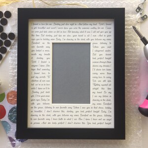 May include: A black picture frame with a white mat and a handwritten love letter inside. The letter is written in cursive script and includes the lyrics to a song. The frame is sitting on a white surface.