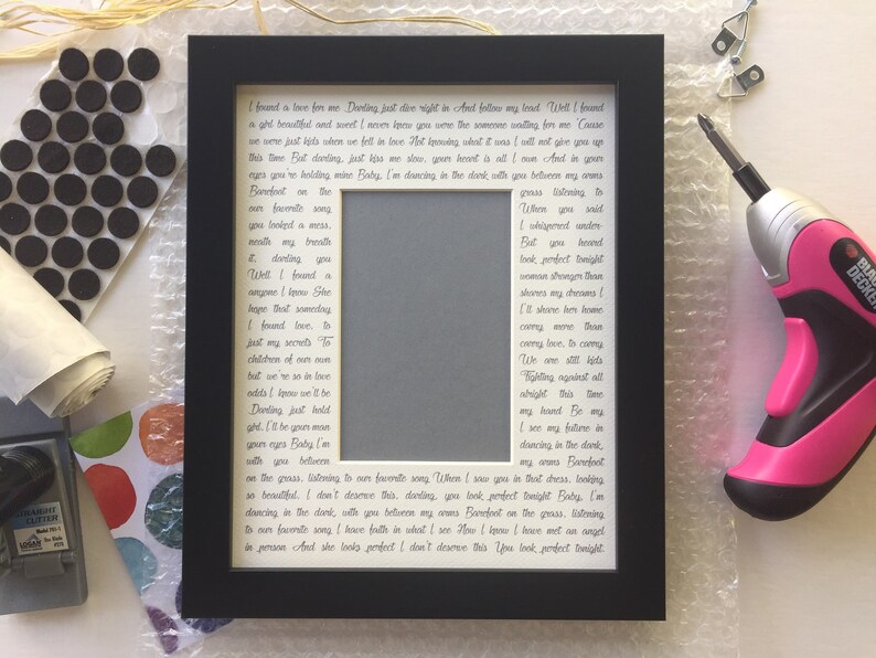 May include: A black picture frame with a white mat and a handwritten love letter inside. The letter is written in cursive script and includes the lyrics to a song. The frame is sitting on a white surface.