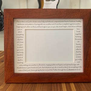 May include: A framed print with a white background and brown text. The text is a bible verse from Colossians 3:12-17. The frame is made of dark wood.