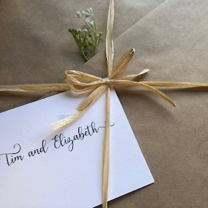 Puede incluir: Una bolsa de regalo de papel marr&oacute;n atada con una cinta de rafia natural y un peque&ntilde;o ramo de flores blancas. La bolsa tiene una tarjeta blanca adjunta con el texto "Tim y Elizabeth".