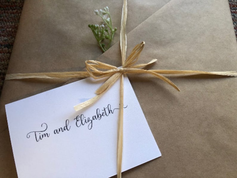 Puede incluir: Una bolsa de regalo de papel marr&oacute;n atada con una cinta de rafia natural y un peque&ntilde;o ramo de flores blancas. La bolsa tiene una tarjeta blanca adjunta con el texto "Tim y Elizabeth".