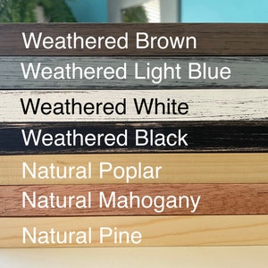 May include: A close-up of seven different wood frame samples. The frames are arranged vertically, with the top frame being weathered brown, followed by weathered light blue, weathered white, weathered black, natural poplar, natural mahogany, and natural pine.