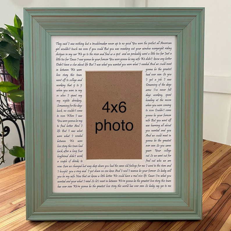 Op de afbeelding: Een 4x6 fotolijst met een lichtblauwe lijst en een bruine passe-partout. De passe-partout heeft een gedrukte tekst over een liefdesverhaal. De tekst bevat de woorden "love story", "college", "job", "dreaming", "forever", "greatest", "ring", "wife", "real nice life", "greatest love story".