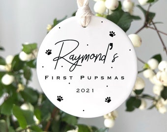 May include: White ceramic Christmas ornament with black paw prints and the text "Raymond's First PupSmas 2021".