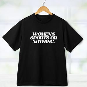 Może przedstawiać: Czarny t-shirt z białym napisem "WOMEN'S SPORTS OR NOTHING." Koszulka wisi na drewnianym wieszaku. Tło to okno z białymi ramami.
