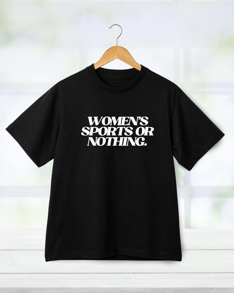 Może przedstawiać: Czarny t-shirt z białym napisem "WOMEN'S SPORTS OR NOTHING." Koszulka wisi na drewnianym wieszaku. Tło to okno z białymi ramami.