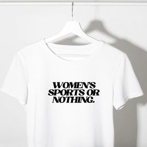 Może przedstawiać: Biały t-shirt z napisem "WOMEN'S SPORTS OR NOTHING" pogrubionymi, czarnymi literami. Koszulka wisi na białym wieszaku na gładkim białym tle. T-shirt ma okrągły dekolt i kr&oacute;tkie rękawy.