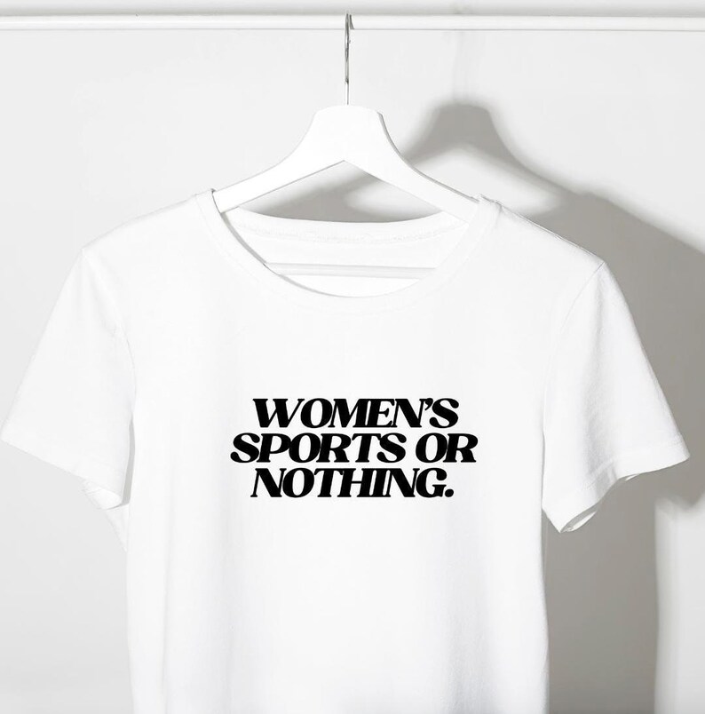 Może przedstawiać: Biały t-shirt z napisem "WOMEN'S SPORTS OR NOTHING" pogrubionymi, czarnymi literami. Koszulka wisi na białym wieszaku na gładkim białym tle. T-shirt ma okrągły dekolt i kr&oacute;tkie rękawy.
