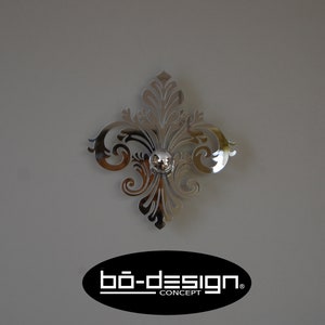 Puede incluir: Un acento de pared decorativo plateado con un dise&ntilde;o floral. El acento tiene un peque&ntilde;o pomo redondo plateado en el centro. El acento est&aacute; montado en una pared gris. El texto "b&ouml;-design. CONCEPT" es visible en la esquina inferior derecha de la imagen.