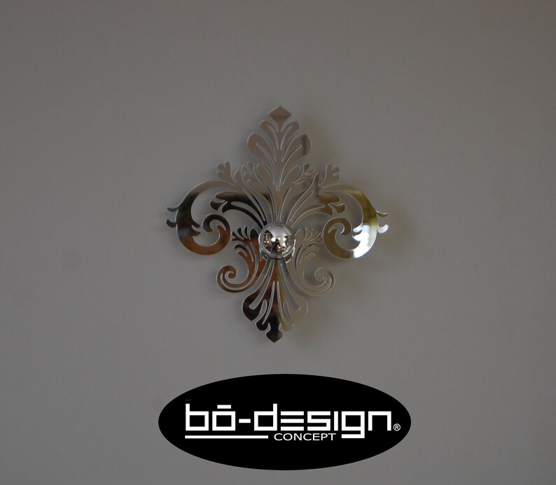 Puede incluir: Un acento de pared decorativo plateado con un dise&ntilde;o floral. El acento tiene un peque&ntilde;o pomo redondo plateado en el centro. El acento est&aacute; montado en una pared gris. El texto "b&ouml;-design. CONCEPT" es visible en la esquina inferior derecha de la imagen.