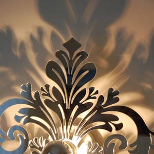 Puede incluir: Una l&aacute;mpara de pared de metal plateado con un dise&ntilde;o floral decorativo. La l&aacute;mpara proyecta una sombra en la pared detr&aacute;s de ella.