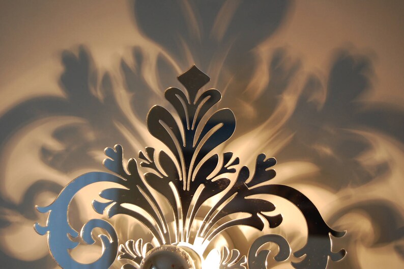 Puede incluir: Una l&aacute;mpara de pared de metal plateado con un dise&ntilde;o floral decorativo. La l&aacute;mpara proyecta una sombra en la pared detr&aacute;s de ella.
