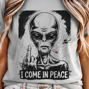 Puede incluir: Camiseta gris claro con un gráfico en blanco y negro de un extraterrestre fumando un cigarrillo y mostrando el dedo medio. El texto "I COME IN PEACE" está impreso debajo de la ilustración del extraterrestre.