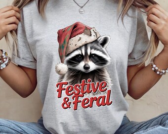 Può includere: T-shirt grigio chiaro con un procione con un cappello di Babbo Natale. Il procione ha la pelliccia bianca e nera ed è centrato sulla maglietta. Il testo "Festive & Feral" è in corsivo rosso sotto.