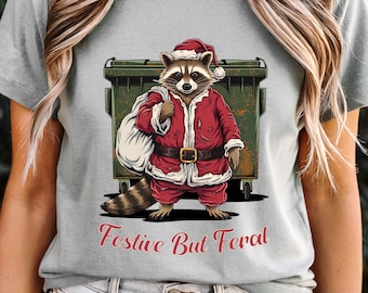 May include: A light grey t-shirt with a raccoon in a Santa Claus outfit, standing before a green skip. The raccoon is in a red Santa suit and hat, carrying a sack. The text "Festive But Feral" is printed in red.