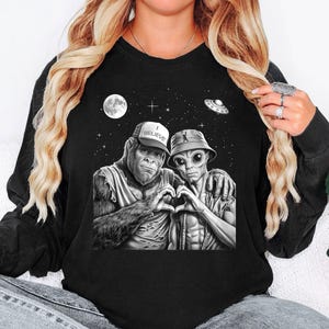 May include: Black sweatshirt with a graphic of a gorilla and an alien forming a heart shape with their hands. The gorilla wears a baseball cap with "I BELIEVE". The alien wears a bucket hat. A moon, stars, and a UFO are in the background.