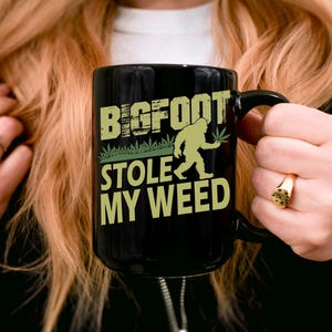 May include: Black coffee mug with a white graphic of a Bigfoot holding a marijuana leaf. The text reads "Bigfoot Stole My Weed".