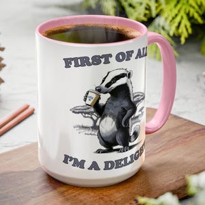 May include: White ceramic mug with a pink rim and handle. The mug features a cartoon badger holding a cup of coffee and the text "First of all I'm a delight".