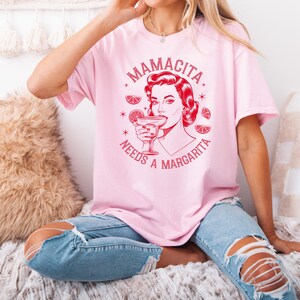 Op de afbeelding: Roze T-shirt met een rode afbeelding van een vrouw die een margarita drinkt met de tekst "Mamacita Needs a Margarita".