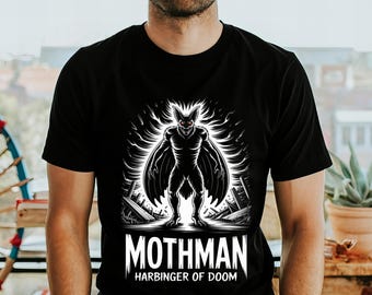 May include: Black t-shirt featuring a detailed, black and white illustration of the Mothman, a cryptid with bat-like wings and glowing red eyes. The text "MOTHMAN HARBINGER OF DOOM" is printed below the image. The shirt is a crew neck.