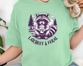 Op de afbeelding: Een lichtgroen T-shirt met een afbeelding van een wasbeer die een kopje koffie vasthoudt. De wasbeer is omringd door bomen en de tekst "Latchkey & Feral" is onder de wasbeer gedrukt.