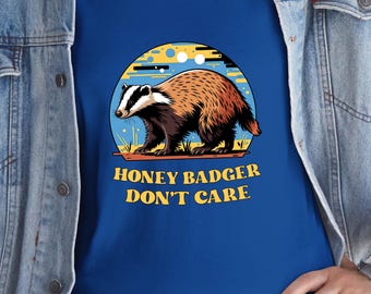 Pode incluir: Uma camiseta azul com uma ilustração de desenho animado de um texugo-do-mel. O texugo é marrom, preto e branco, em um fundo amarelo e azul. O texto "HONEY BADGER DON'T CARE" é amarelo.