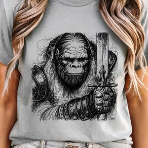 Puede incluir: Camiseta gris claro con una ilustración en blanco y negro de un gorila guerrero sosteniendo una espada. El gorila lleva armadura y tiene una expresión seria. La obra de arte es detallada y la camiseta es de color sólido.