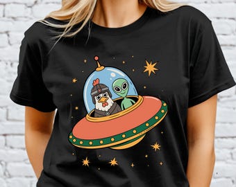 May include: Black t-shirt featuring a cartoon chicken in a helmet and a green alien in a red and orange spaceship. The spaceship is surrounded by stars. The design is whimsical and fun.