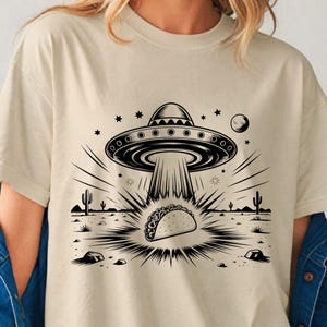 Könnte beinhalten: Beiges T-Shirt mit einer schwarz-weißen Grafik eines UFOs, das einen Taco entführt. Das Design umfasst Sterne, einen Mond, Kakteen und eine Wüstenlandschaft. Das T-Shirt hat kurze Ärmel und einen Rundhalsausschnitt.