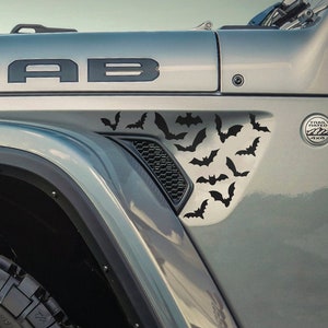 May include: A gray Jeep Wrangler with a black "MOAB" decal on the hood. The vehicle has a black bat decal on the side of the fender.