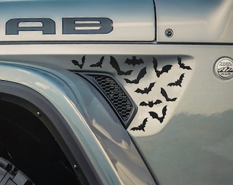 Könnte beinhalten: Ein grauer Jeep Wrangler mit einem schwarzen "MOAB"-Aufkleber auf der Motorhaube. Das Fahrzeug hat einen schwarzen Fledermaus-Aufkleber an der Seite des Kotflügels.