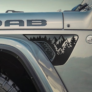 May include: A gray Jeep Wrangler with a black "MOAB" decal on the hood and a black mountain scene decal on the fender. The Jeep has a black tire and a black fender flare.