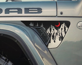 Puede incluir: Un Jeep Wrangler gris con una calcomanía negra "MOAB" en el capó. El vehículo tiene una calcomanía de escena de montaña en blanco y negro en el guardabarros con una luna creciente roja.
