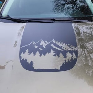 May include: A car hood with a dark gray decal featuring a mountain range and pine trees. The decal is a silhouette design, with the mountains and trees in a lighter gray color. The car hood is a light olive green color.