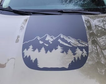 May include: A car hood with a dark gray decal featuring a mountain range and pine trees. The decal is a silhouette design, with the mountains and trees in a lighter gray color. The car hood is a light olive green color.