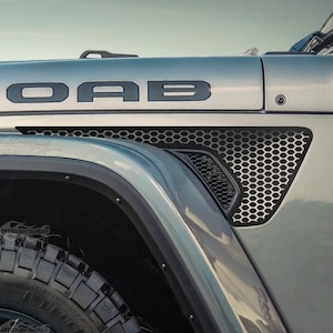 May include: A silver vehicle side panel featuring a black MOAB decal on the hood, a black honeycomb-patterned vent insert, and a circular Trail Rated 4x4 badge. The vehicle has a black wheel arch trim and a black tire with a rugged tread pattern.