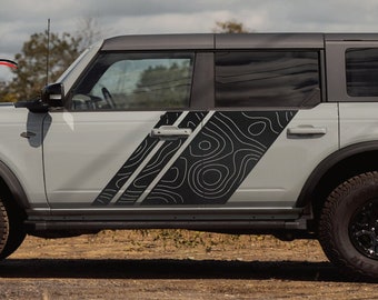 May include: A gray Ford Bronco with black and gray topographic stripe decals on the side. The vehicle is parked in a field with tall grass.
