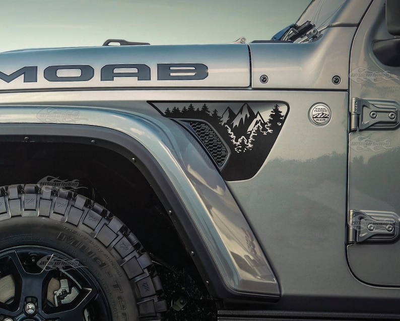 May include: A gray Jeep Wrangler with a black "MOAB" decal on the hood and a black mountain scene decal on the fender. The Jeep has a black tire and a black fender flare.